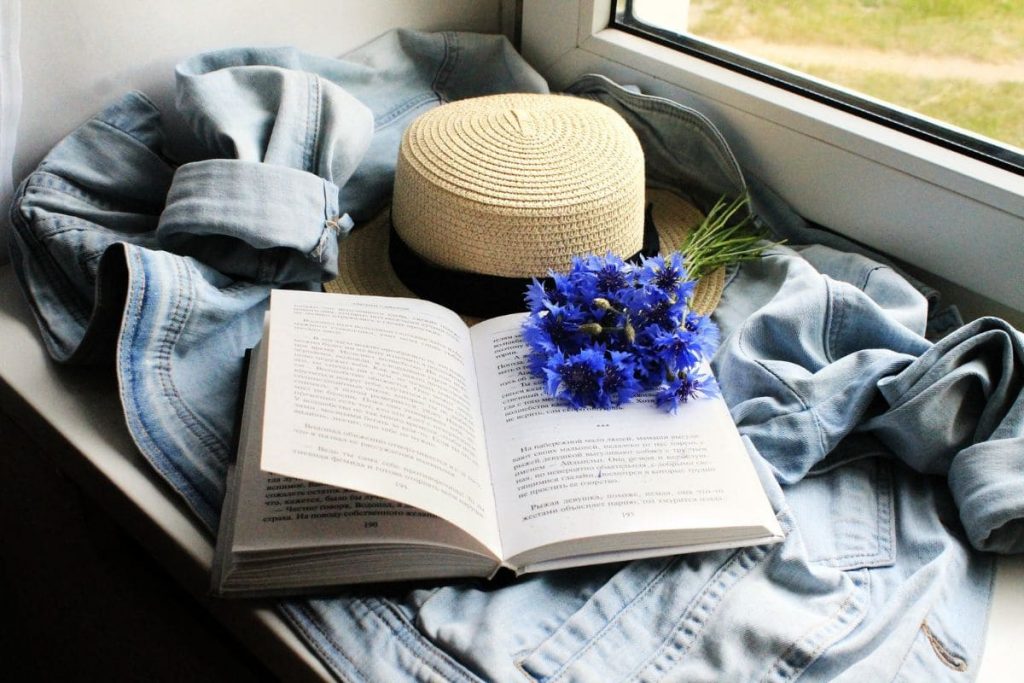 Open book with blue flowers. A hat and a jeans shirt by window.