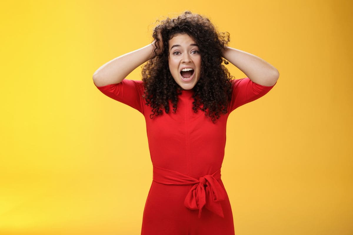 Girl feeling pressure, standing anxious in panic, holding hands on curly black hair, yelling at camera. Disturbed, freaked out being tensed and upset with bad situation, standing troubled over yellow background
