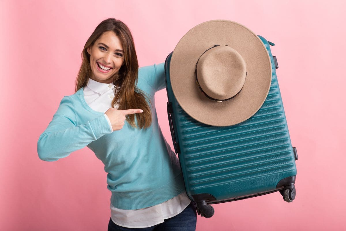 Young woman travelling alone, pointing on her suitcase on pink background